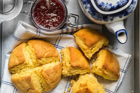 Gyllene scones majsmjöl, en burk hallonsylt och en blåvit tekanna. Perfekt för en mysig fika.