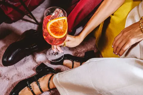 Elegancka dłoń trzyma kieliszek z drinkiem, w którym króluje wytrawny wermut. Obok błyszczą czarne buty i złota biżuteria.