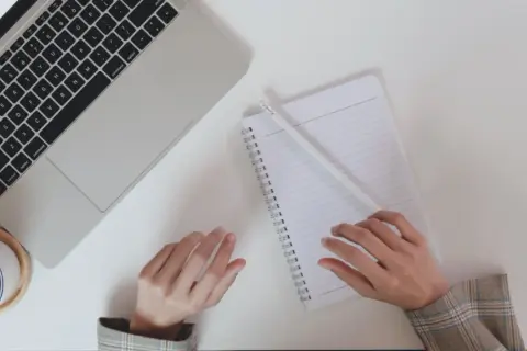 Dłonie nad notatnikiem i laptopem, filiżanka kawy.