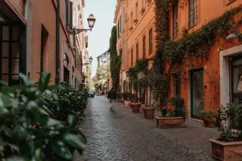 Eine Person geht durch eine schmale Gasse in Rom, gesäumt von orangefarbenen Gebäuden und Pflanzen. Ein perfekter Ort für Rom Geheimtipps.