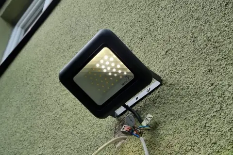 Czarny reflektor LED zamontowany na zielonej ścianie. Żarzące się ledy pobierają prąd, co widać po podłączonych przewodach.