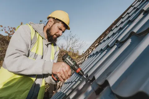 A roofer in a yellow hard hat and hi-vis vest uses a drill for metal roof repair.