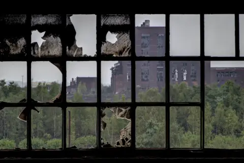 Through a broken window pane, a scene of urbex photography unfolds: decaying brick buildings stand amidst overgrown trees, a testament to time's passage.