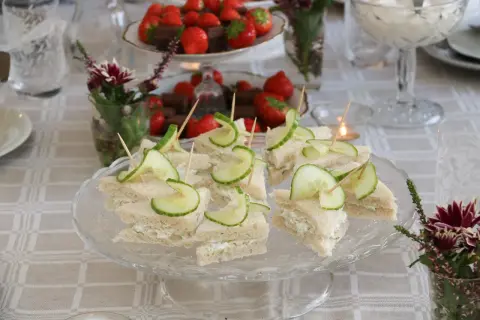 Små smörgåsar med gurka och gräddost, redo för afternoon tea. Jordgubbar och choklad i bakgrunden.
