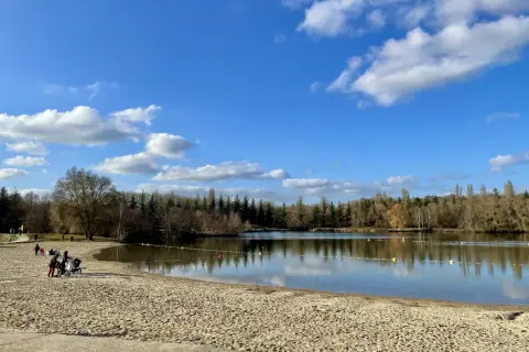 Plage de sable fin au bord d'un lac paisible, avec des familles profitant du soleil. La **cascade** de la **forêt de Fontainebleau** se reflète dans le **lac** sous un ciel bleu parsemé de nuages.