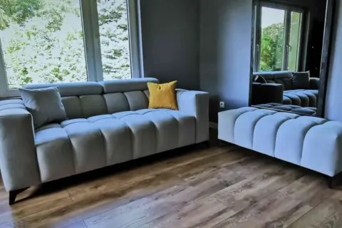 Nowoczesna szara sofa i pufa, ozdobione żółtą poduszką. To meble od najlepszych producentów mebli tapicerowanych, idealne do stylowego salonu.