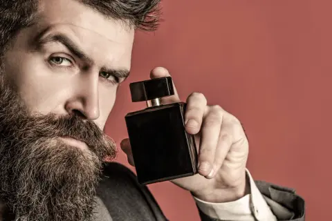 Mężczyzna z brodą trzyma czarną buteleczkę perfum na czerwonym tle.