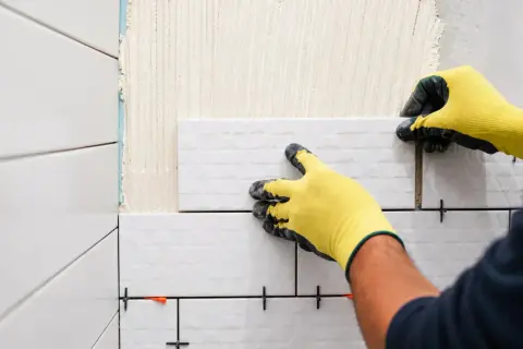 Mains gantées jaunes en pleine pose carrelage mural blanc. Des croisillons maintiennent l'espacement.