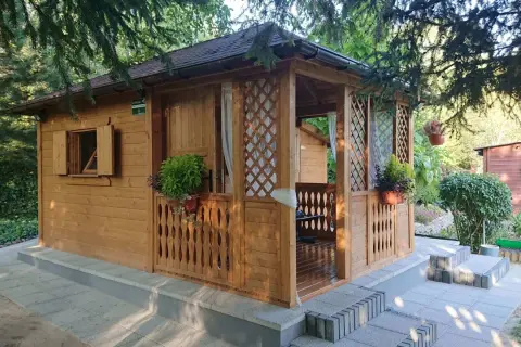 Drewniany domek letniskowy z werandą, otoczony zielenią. Czy domek letniskowy można przekształcić na mieszkalny? Ten wygląda na gotowy do adaptacji.