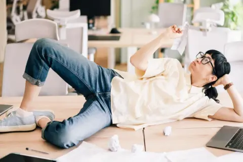 En person ligger utsträckt på ett skrivbord, omgiven av papper och laptops. Klädd i jeans och en ljus skjorta, ser hen ut att fundera över en papperslapp, en bild av en dysfunktionell arbetsplats.