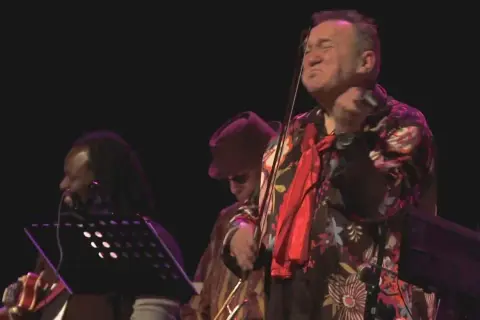 Michał Olczak gra na skrzypcach, ubrany w kolorową koszulę. Obok niego muzycy z dredami i w kapeluszu.