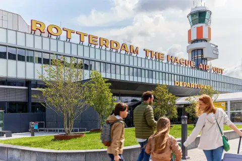 Rodzina zmierza na lotnisko Rotterdam The Hague Airport. W tle widoczna wieża kontroli lotów i napis z nazwą lotniska.