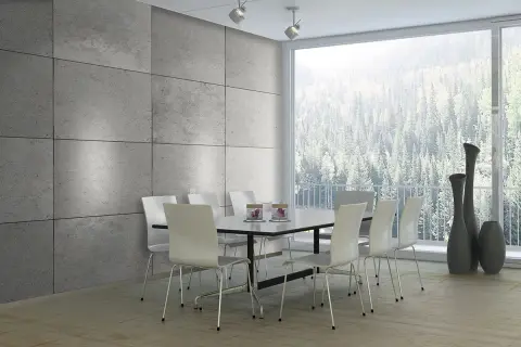 Esszimmer mit moderner Beton Optik Wandverkleidung, weißem Tisch und Stühlen, sowie einem Panoramafenster mit Blick auf einen Wald.