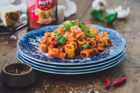 En tallrik med rigatoni all'arrabiata, toppad med basilika och parmesan. Bredvid syns en burk Mutti-tomatsås, vitlök och chilifrukter.