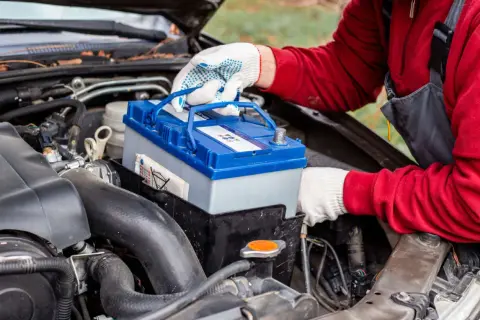 Persona con guantes blancos manipula una batería de coche azul y gris, lista para cargar batería coche.