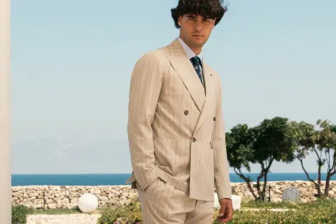 Uomo elegante con abito gessato beige, in piedi con lo sguardo rivolto verso l'osservatore, con il mare e il cielo azzurro sullo sfondo.