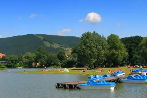 Słoneczny dzień nad jeziorem w Międzybrodziu Żywieckim. Ludzie odpoczywają na plaży, a przy brzegu cumują niebieskie rowerki wodne.