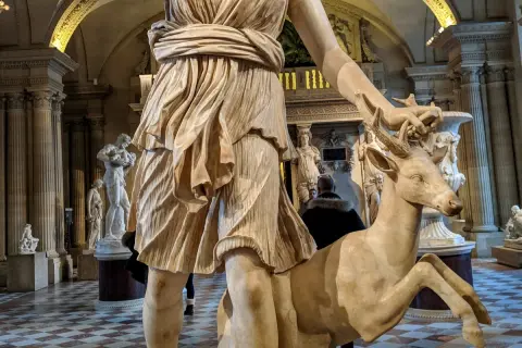 Statue de Diane chasseresse, qui est Artémis, tenant un cerf. L'œuvre est dans un musée, avec d'autres sculptures en arrière-plan.