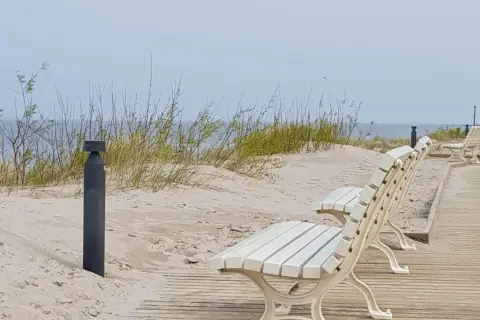 Białe ławki na drewnianej promenadzie w Dziwnowie, z widokiem na piaszczyste wydmy i morze.