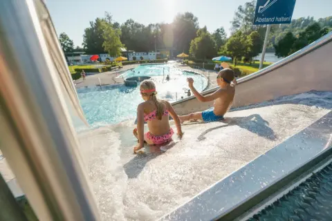 Kinder rutschen im Freibad Leutkirch auf einer Wasserrutsche. Im Hintergrund das Schwimmbecken mit Badegästen.