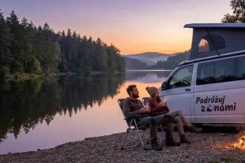 Para relaksuje się przy ognisku podczas **camping al lago**. W tle spokojne jezioro i las o zachodzie słońca.