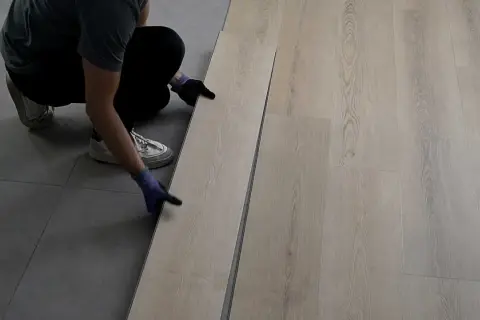 Persona con guantes instala piso SPC de madera clara sobre suelo gris.