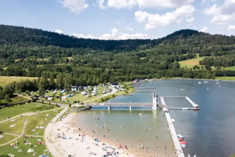 Słoneczny dzień nad zalewem Stara Morawa. Ludzie odpoczywają na plaży i w wodzie, a w tle widać las i pola namiotowe.