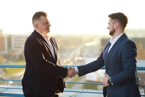 Dwóch mężczyzn w garniturach ściska sobie dłonie na tle miasta.