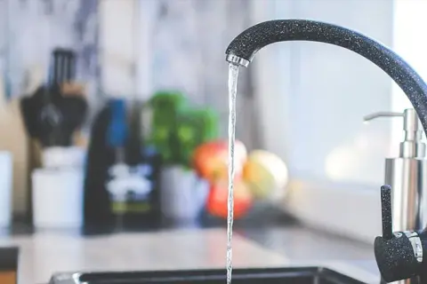 Un robinet moderne coule dans une cuisine. Un préparateur ECS discret est visible à l'arrière-plan, à côté d'un distributeur de savon.