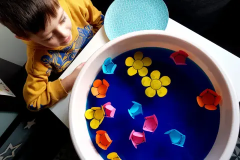 Chłopiec w żółtej bluzie obserwuje kolorowe papierowe kwiatki unoszące się na wodzie w misce.