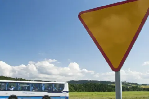 Żółty trójkątny znak a7 ostrzega przed ustąpieniem pierwszeństwa. W tle jedzie autobus, a za nim zielone wzgórza.
