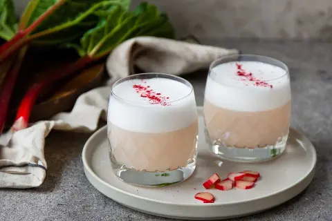 Två glas med krämig rabarber sockerlag, toppade med skum och strössel. Färska rabarberstjälkar i bakgrunden.
