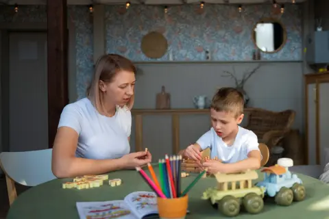 Kobieta i chłopiec grają w drewniane gry na zielonym stole. W tle widać wystrój pokoju, przypominający salę w przedszkolu niepublicznym.