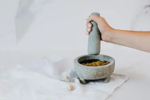 Dłoń uciera składniki w moździerzu, pokazując, jak zrobić pesto. Obok leżą ząbki czosnku.