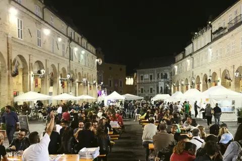 Serata di festa in una piazza delle Marche, con tavoli all'aperto e gente che mangia e socializza.