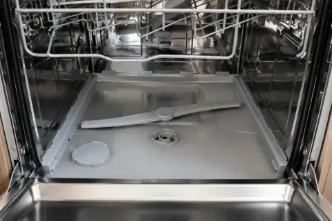 Intérieur d'un lave-vaisselle ouvert, montrant le bras d'aspersion et le fond. On observe de l'eau stagnante au niveau du filtre, signe d'un nettoyage nécessaire.
