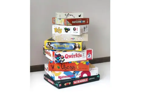 Stos pudeł z grami planszowymi, idealnymi na rodzinne wieczory. Wśród nich "Splendor", "Qwirkle", "Ubongo" i "Scrabble" – świetna gra dla seniorów.