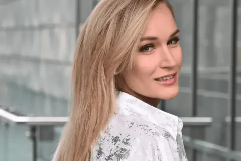 Uśmiechnięta Kamila Boś, w białej koszuli z wzorem, patrzy przez ramię.