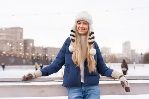 Uśmiechnięta blondynka w granatowej kurtce i białej, futrzanej czapce z pomponami. Idealny zestaw na lodowisko.