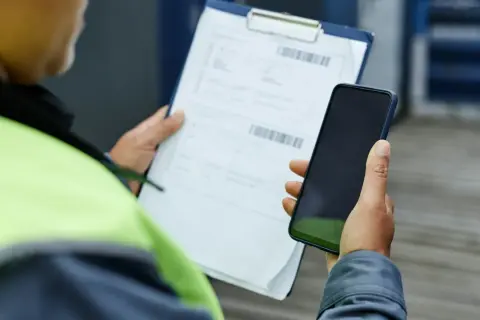 Pracownik w kamizelce odblaskowej trzyma telefon i dokumenty. Zastanawia się, czy warto złożyć skargę do PIP.