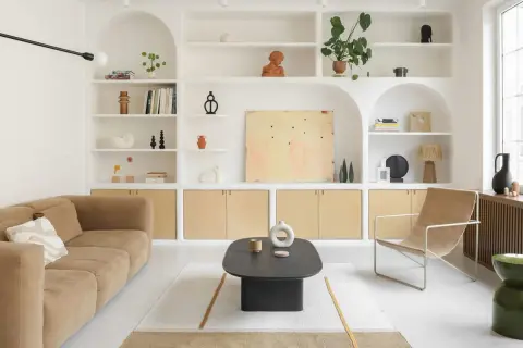 Salon épuré avec canapé beige, fauteuil en toile, table basse noire et étagères murales remplies d'objets décoratifs. Le tout baigne dans une douce lumière, rehaussée par la **couleur blanc cassé** des murs et des étagères.