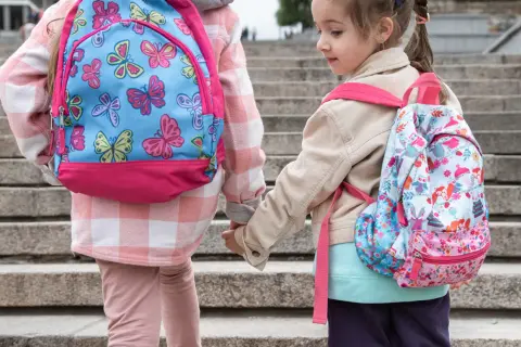Dwie dziewczynki w kolorowych plecakach szkolnych trzymają się za ręce na schodach.