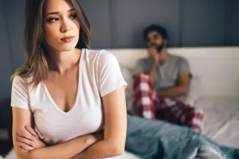 Eine Frau blickt nachdenklich weg, während ein Mann im Hintergrund sitzt. Die Stimmung ist angespannt, ein Zeichen, wann ist eine beziehung nicht mehr zu retten.