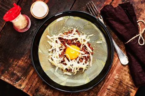 En läcker koreansk råbiff med en gul äggula i mitten, toppad med strimlad rättika och sesamfrön. Serveras på en vacker tallrik med en mörk servett och gaffel.