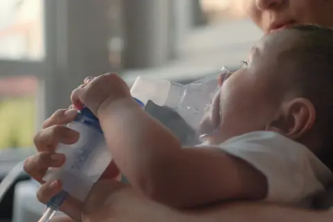 Mama pomaga maluszkowi z mokrym kaszlem u niemowlaka, stosując inhalator.