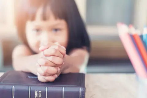Dziecko z modlącymi się dłońmi na Biblii, obok kolorowe kredki. Nauka 10 przykazań bożych dla dzieci.