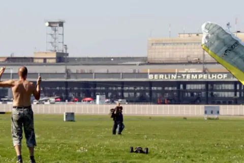 Ein Mann mit nacktem Oberkörper steht auf dem Feld vor dem BERLIN-TEMPELHOF. Ein Gleitschirm liegt im Vordergrund.