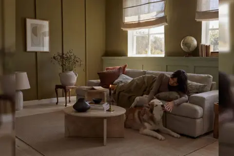 Gemütliches Wohnzimmer in warmen Farben. Eine Frau kuschelt mit ihrem Hund auf dem Sofa, umgeben von erdigen Tönen und sanftem Licht.