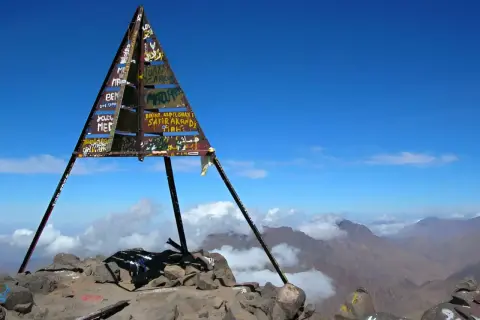 Sommet du mont Toubkal, altitude impressionnante. Le cairn triangulaire, couvert de graffitis, se dresse fièrement sur un pic rocheux, dominant un paysage montagneux spectaculaire sous un ciel bleu éclatant.