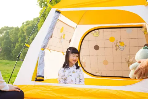 Rodzina rozkłada żółty namiot plażowy. Uśmiechnięta dziewczynka w środku, rodzice pomagają.